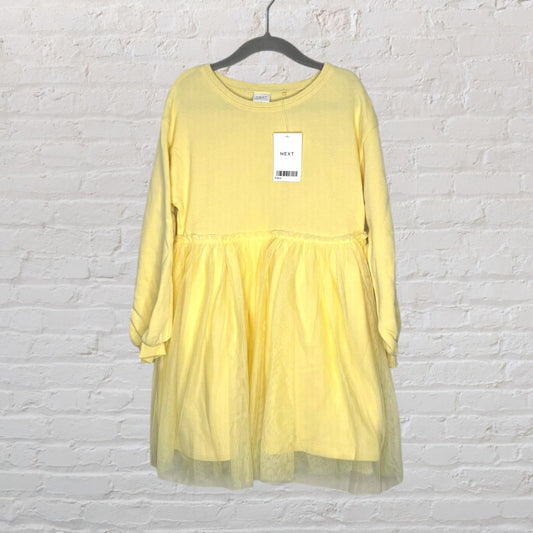 Yellow long sleeve tulle dress with a tag on a hanger, by Next, against a white brick wall.