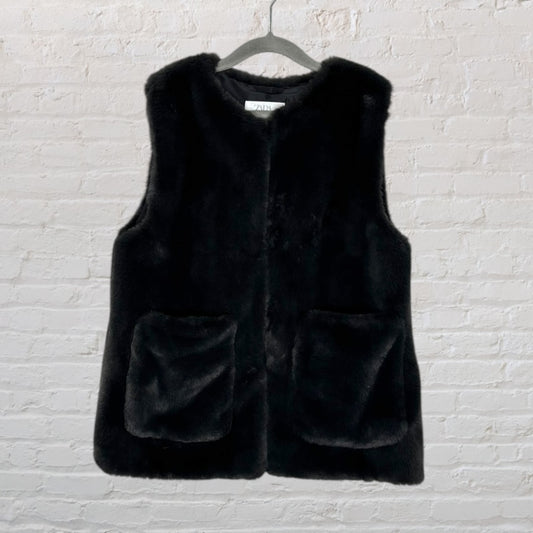 Black faux fur sleeveless vest by Zara on a hanger against a white brick wall.