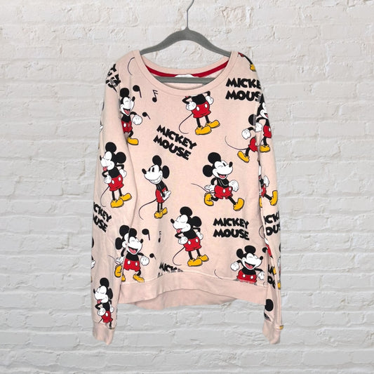 Pink sweatshirt featuring a repeated print of Mickey Mouse in different poses, hanging on a hanger against a white brick wall.