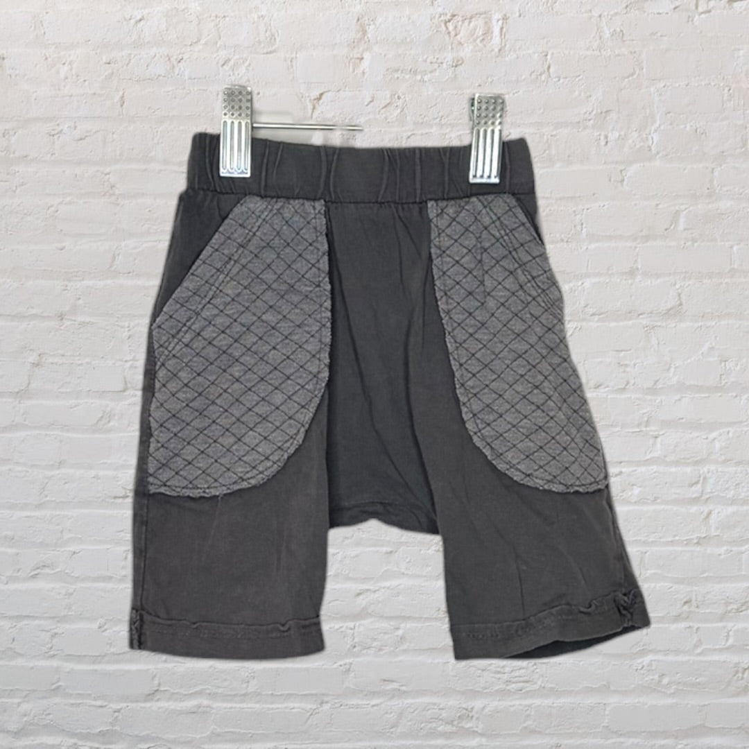 Dark grey shorts with quilted pockets, hanging on a hanger against a white brick wall.