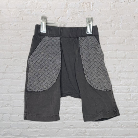 Dark grey shorts with quilted pockets, hanging on a hanger against a white brick wall.