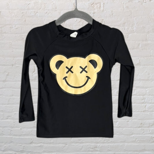 Black long sleeve shirt with a large yellow smiley face bear graphic, hanging on a hanger against a white brick wall.
