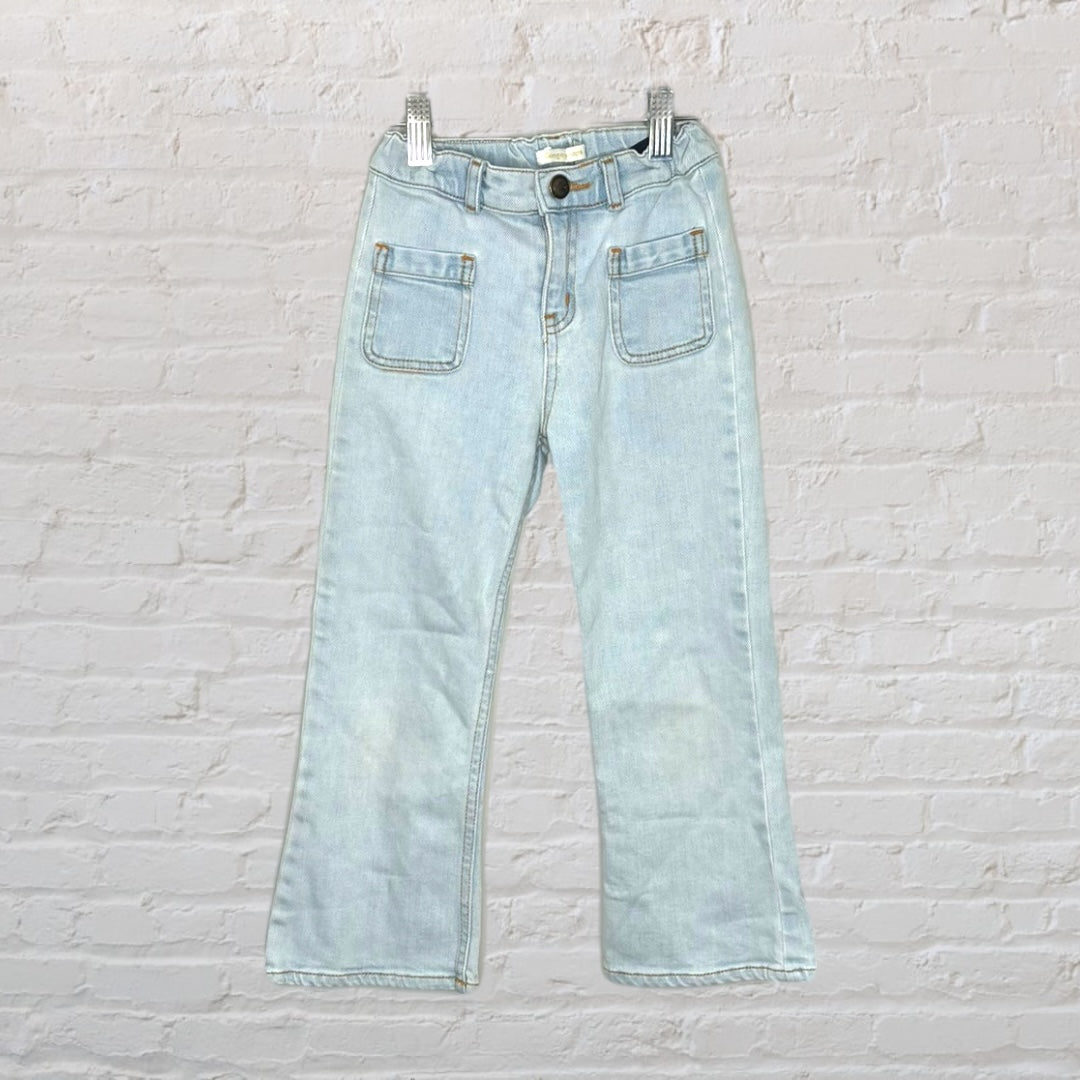 Light wash wide-leg denim jeans with front patch pockets, displayed hanging against a white brick wall.