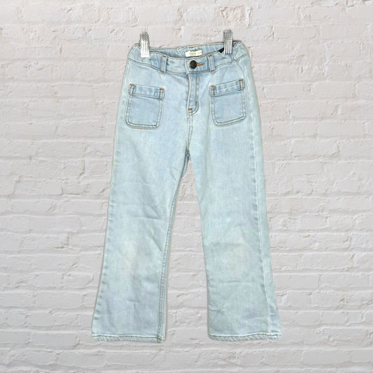Light wash wide-leg denim jeans with front patch pockets, displayed hanging against a white brick wall.