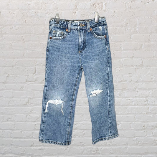 Blue denim jeans with distressed rips displayed against a white brick wall.