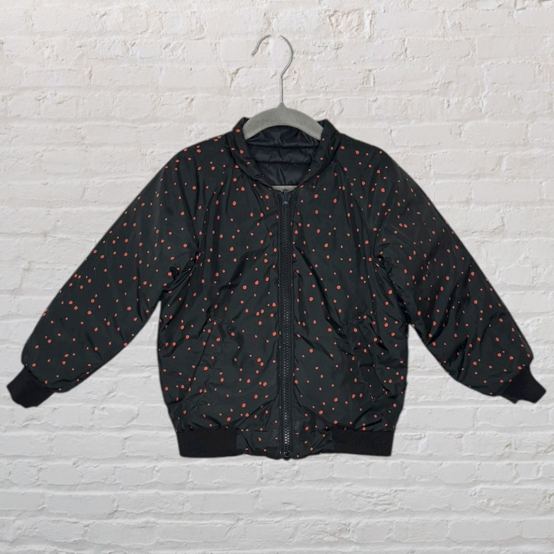 Black puffer jacket with red polka dots, hung on a hanger against a white brick wall.