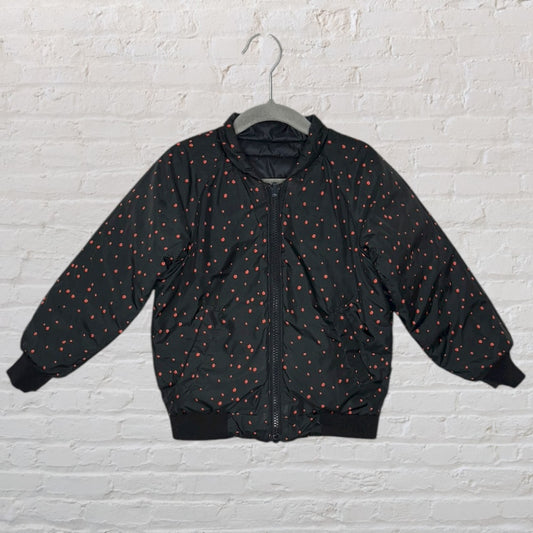 Black puffer jacket with red polka dots, hung on a hanger against a white brick wall.