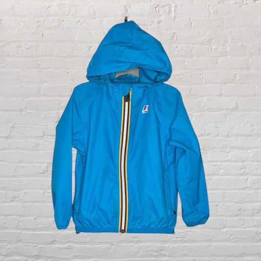 Blue K-Way hooded windbreaker jacket with a multicolored zipper, displayed on a hanger against a white brick wall.