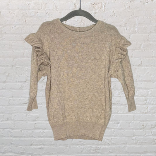 Beige cable knit sweater with ruffled details on the shoulders, hanging on a black hanger against a brick wall.