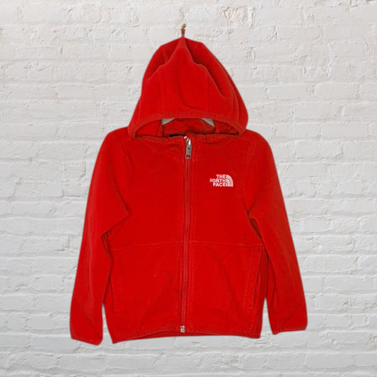 Bright red fleece zip-up hoodie by The North Face with a hood, displayed against a white brick wall.