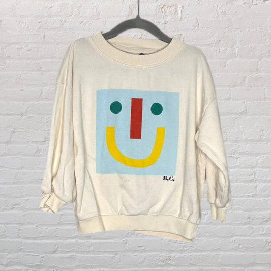 Cream-colored sweatshirt with a colorful geometric smiley face graphic and 'B.C.' initials on the front.