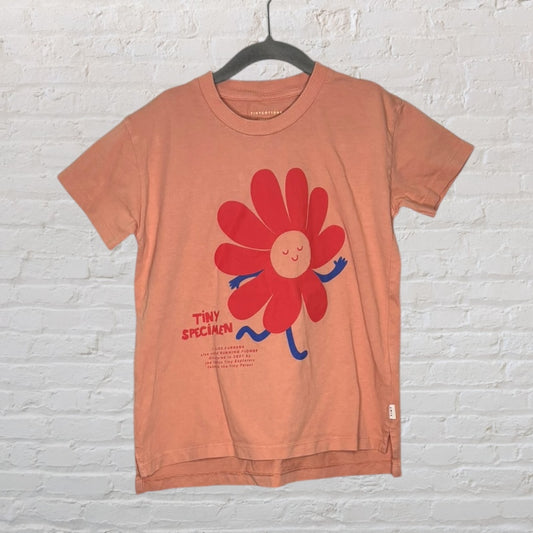 Peach T-shirt featuring a red flower with a smiling face and 'Tiny Specimen' text, hanging on a black hanger.