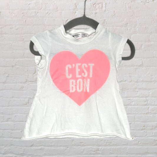White t-shirt with a pink heart and 'C'est Bon' text, by Noah Love, hanging on a black hanger.