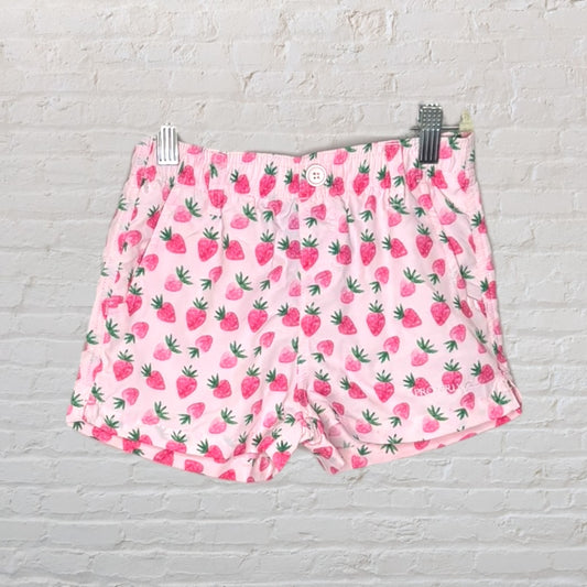 Pink shorts with a strawberry print pattern featuring green leaves, displayed on a hanger against a brick wall background.