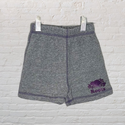 Grey knit shorts with a purple Roots logo, displayed against a white brick background.