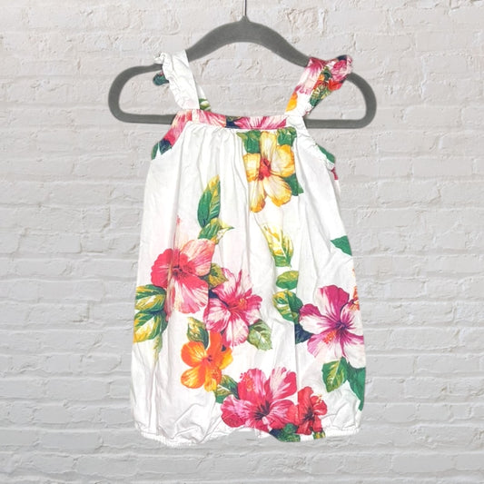 White sundress with colorful floral print, featuring ruffled straps, hanging on a dark hanger against a white brick wall.