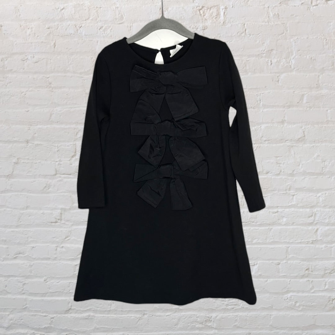 Elegant black dress with bow embellishments down the front, featuring long sleeves and rounded neckline, hanging on a gray hanger.