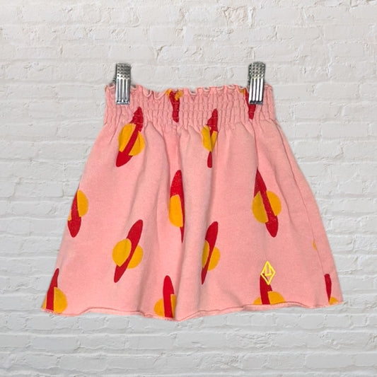 Pink skirt with red and yellow planet pattern and elastic waistband, displayed on hangers against a white brick wall.