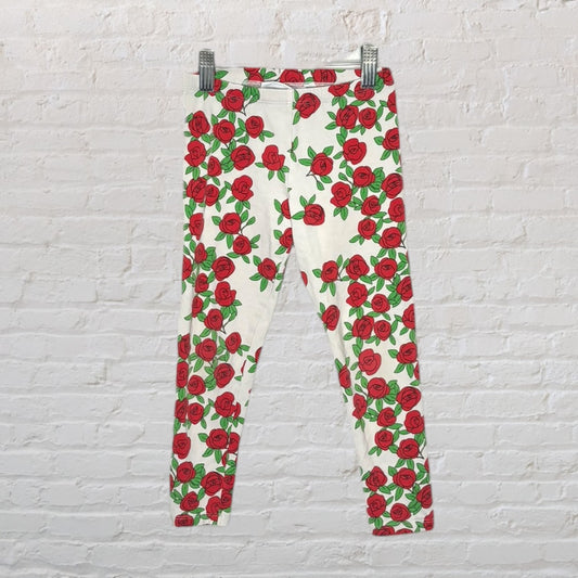 White leggings with a vibrant red rose print pattern throughout, hanging against a white brick wall.