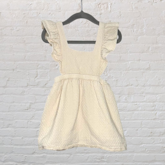 Cream-colored pinafore dress with ruffled sleeves and a textured fabric, hanging on a black hanger.