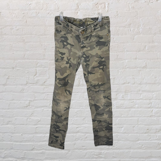 Camouflage print skinny pants hanging against a white brick background.